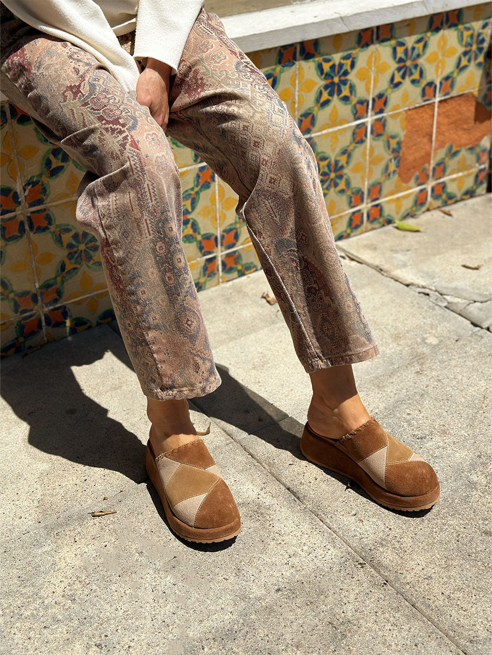 Person wearing patterned pants and brown shoes against a colorful tiled wall.